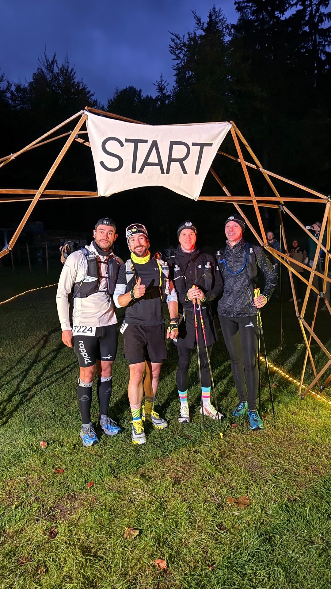 Vier Läufer mit Stirnlampen und Laufwesten posieren kurz vor dem Start unter dem hölzernen Startbogen mit START-Banner